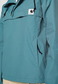 Niebieska nylonowa kurtka z kieszenią na froncie, regulowanymi sznurkami i naszywką z logo na klatce piersiowej. Gładka tekstura i luźny krój.