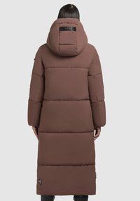 Barna puffernadrág kapucnival, átöltöztetett dizájnnal és hátsó logóval. Hosszú, laza szabású, oldalsó zsebekkel.