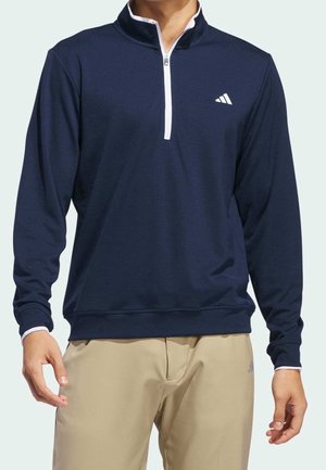 Mann trägt einen marineblauen Langarm-Quarter-Zip-Pullover mit weißem Besatz und beigen Hosen, steht mit locker an den Seiten hängenden Händen.