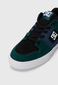 Grüne und schwarze Wildleder-Sneaker mit einem marineblauen Einsatz, schwarzen Schnürsenkeln, einer weißen Gummisohle und einem auffälligen DC-Logo an der Seite.