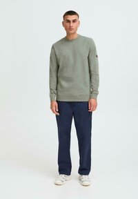 Grønn genser med rund hals, teksturert stoff og en brun detalj på ermene, kombinert med marineblå bukser og hvite sko.