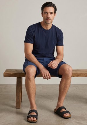 Mand med mørkt hår iført marineblå t-shirt, blå shorts og sorte sandaler, siddende på en træbænk mod en ensfarvet baggrund.