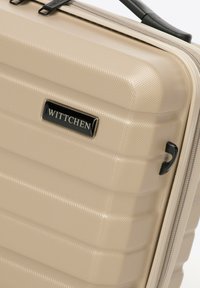 Valigia rigida di colore beige chiaro con superficie testurizzata, caratterizzata da scanalature verticali, una placca con logo nera e una zip.