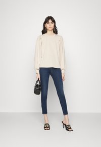 Beige dlouhý rukáv s nabíranými ramenními švy, zkombinovaný s tmavě modrými skinny džíny a černými sandály na podpatku, v ruce držící malou černou kabelku.