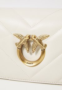 Κρέμα δερμάτινη θήκη με σχέδιο διαγωνίων επενδύσεων, που διαθέτει κούμπωμα με τραπεζοειδές πουλί και γλυπτό δαχτυλίδι. Μινιμαλιστικός σχεδιασμός και απαλός ιστός.