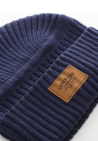 Beanie lavorato a maglia di colore blu navy con texture a costine, dotato di una patch in pelle tan etichettata "Timberland Company". Materiale robusto e caldo.