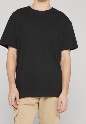 Homme portant un T-shirt noir uni à manches courtes et un pantalon cargo beige, debout devant un fond neutre.