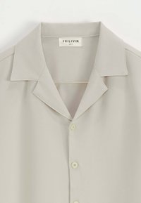 Chemise beige clair à manches courtes avec un col, comportant cinq boutons, une texture lisse et une étiquette portant la mention "FRILIVIN TAILLE 5."