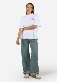 T-shirt surdimensionné blanc avec un logo circulaire rouge, associé à un jean ample vert clair et des baskets beiges avec un motif blanc.