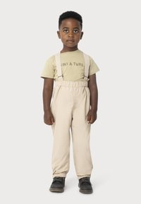 Beige Hosenträger über einem hellgrünen Kurzarmhemd mit dem Text "UNI À TURE". Das Kind trägt schwarze Schuhe und hat lockiges Haar.