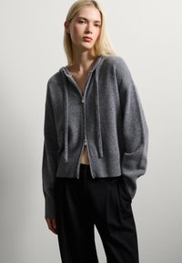 Felpa grigia con zip, realizzata in un morbido e caldo tessuto, con una vestibilità comoda, orlo a costine e lacci sul cappuccio. Abbinata a pantaloni neri.