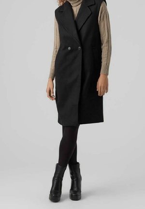 Donna che indossa un cappotto nero senza maniche sopra a un maglione dolcevita beige, leggings neri e stivaletti neri con piattaforma, in piedi davanti a uno sfondo semplice.