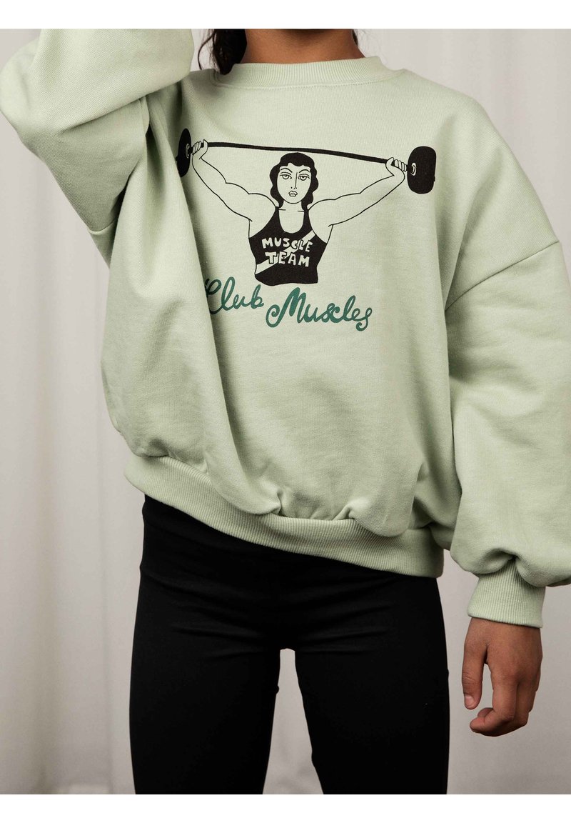 Personne portant un sweat-shirt vert clair avec un graphisme d'une femme musclée soulevant des haltères et le texte "Muscle Team Club Muscles."