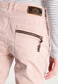 Jasnoróżowe spodnie z teksturowanym akcentem w kieszeni, metalowym zamkiem i drewnianym detalem. Czarna skórzana naszywka z logo na pasku.
