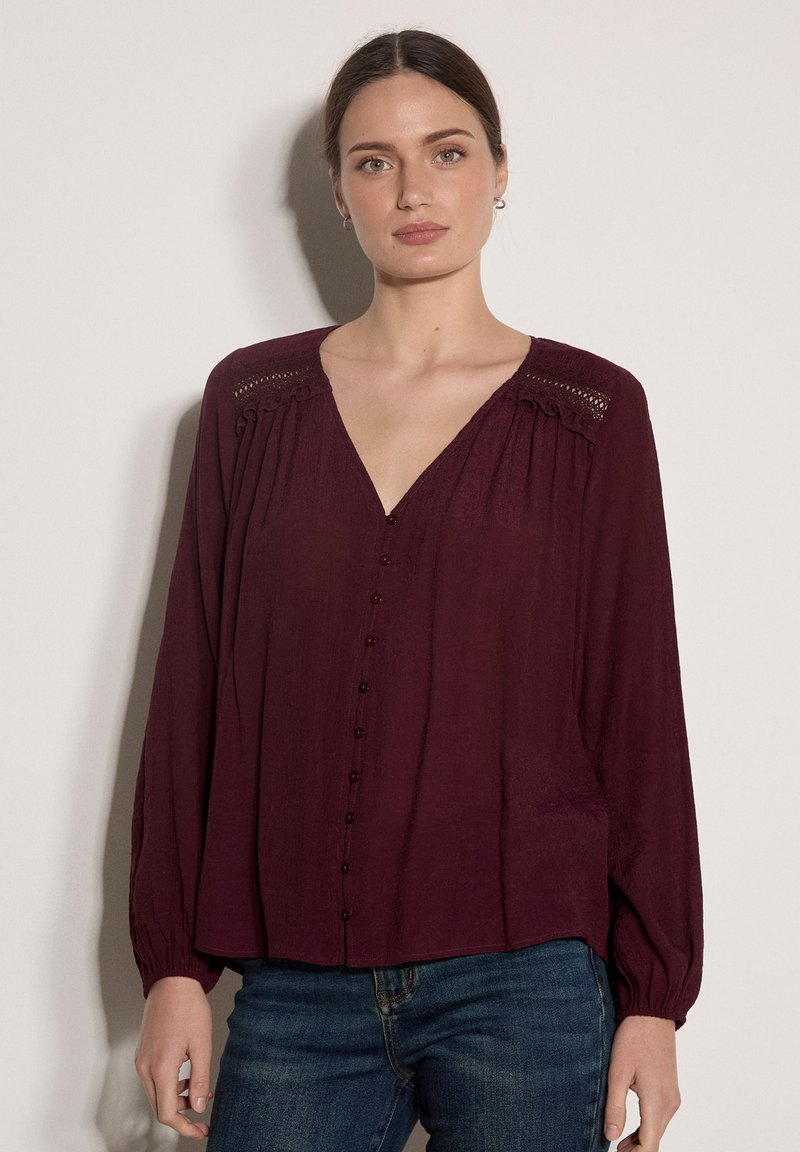 Femme aux cheveux bruns attachés, portant un chemisier rouge foncé à manches longues avec des boutons et un jean bleu, debout devant un fond clair.