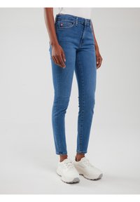 Sieviete valkā pieguļošas zilas džinsa bikses un baltas masīvas sporta kurpes, stāvot pret vienkāršu gaišu fona sienu.