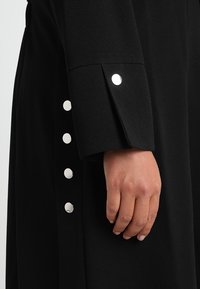 Černý rukáv kabátu s hladkým povrchem, stříbrnými patentkami na manžetách a přiléhavým střihem pro elegantní vzhled.