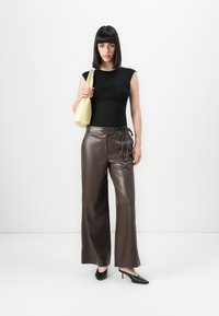 Una donna con capelli scuri e lisci indossa un top senza maniche nero, pantaloni in pelle larghi marroni, décolleté neri e tiene una borsa a spalla gialla pallida.