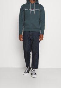 Sudadera con capucha verde oscuro con cordones, texto de logo blanco, bolsillo tipo canguro, combinada con pantalones vaqueros oscuros y zapatillas negras.