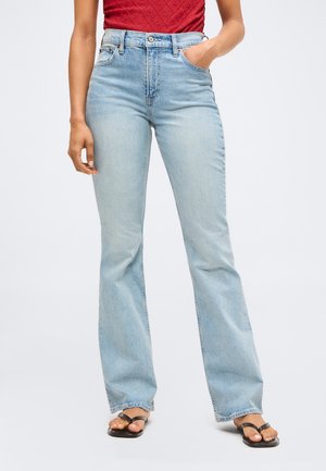 Γυναίκα που φοράει ανοιχτό μπλε τζιν ψηλόμεσο με καμπάνα, κόκκινο αμάνικο τοπ και μαύρα σανδάλια, στέκεται με το ένα χέρι στην τσέπη.