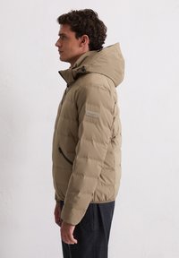 Beige Steppjacke mit durchgehendem Reißverschluss, Kapuze und Seitentaschen. Verfügt über eine gesteppte Textur und ein Etikett am Ärmel. Geeignet für kaltes Wetter.