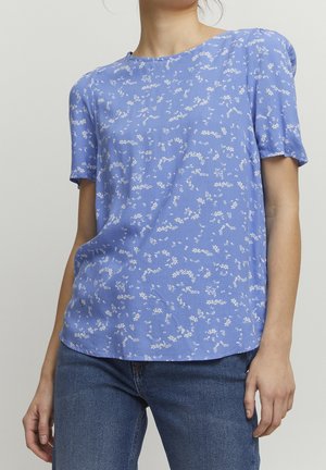 Kvinde iført en lyseblå kortærmet bluse med hvidt blomsterprint og blå jeans, stående foran en ensfarvet baggrund.