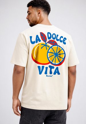 Man wearing off-white t-shirt with colorful orange fruit graphic and text "LA DOLCE VITA" on the back, standing against a plain background.