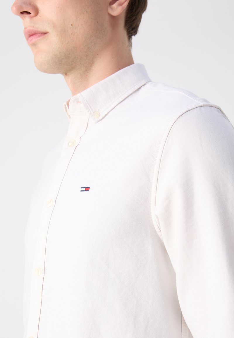 Camisa blanca de botones con cuello, que presenta un pequeño logo bordado en rojo, blanco y azul en el pecho. Textura de tela suave.