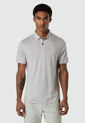 Uomo con capelli corti e ricci, indossa una polo a maniche corte grigio chiaro e pantaloni chiari, in piedi davanti a uno sfondo semplice.