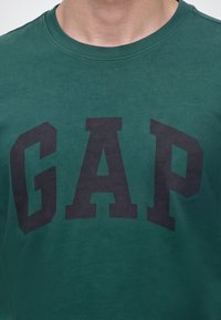 Homme portant un sweat-shirt vert à col rond avec de grandes lettres noires formant le mot GAP sur la poitrine.