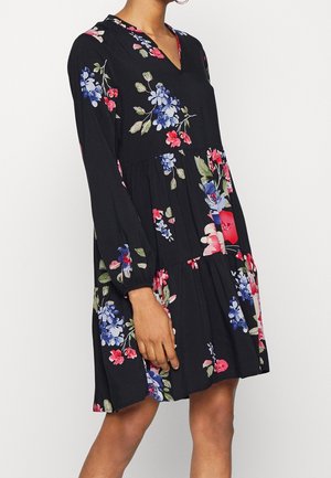 Kobieta ubrana w czarną sukienkę z długim rękawem z niebieskimi, czerwonymi i różowymi kwiatowymi wzorami, stojąca na jednolitym jasnym tle.