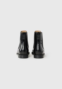 Une paire de bottines en cuir noir brillant avec des panneaux élastiques sur les côtés, vue de l'arrière sur un fond blanc uni.