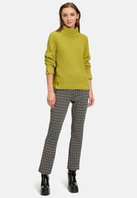 Grüner Strickpullover mit hohem Kragen, kombiniert mit gemusterten schwarz-weißen Hosen. Das Model trägt schwarze Plateastiefel.