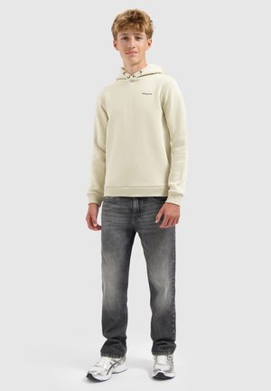 Junger Mann steht in einem hellbeigen Hoodie, ausgewaschenen schwarzen Jeans und silbernen Laufschuhen vor einem schlichten grauen Hintergrund.