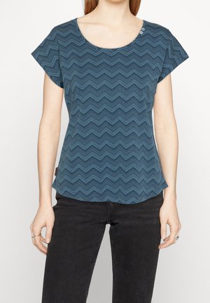 Mujer con camiseta azul de manga corta con estampado de zigzag y vaqueros negros, de pie frente a un fondo liso y claro.