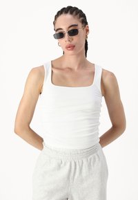 Hvit, tettsittende tank topp med firkantet halslinje og teksturert stoff. Kombinert med grå joggebukser med elastisk midjebånd og sidelommer.