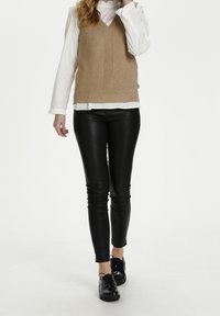 Béžová pletená vesta s výstřihem do V a chevron vzorem, přes bílou košilovou blůzu. Černé kožené legíny a černé lakované šněrovací boty.