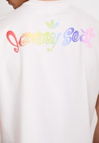 Hvid bomulds-t-shirt med en farverig, stiliseret tekst "Jeremy Scott" hen over ryggen, toppet med et grønt Adidas-logo.