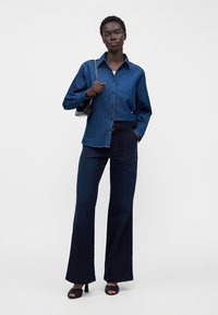 Femme debout portant une chemise en jean bleu foncé, un pantalon large sombre, des talons noirs et un petit sac à main noir avec une bandoulière chaîne.
