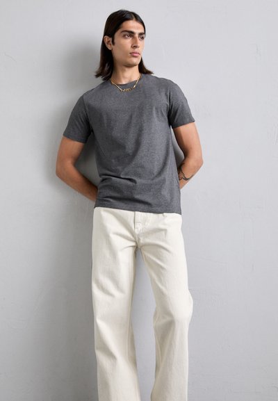 Grå bomull t-skjorte kombinert med kremfargede, rette jeans. Figuren står mot en strukturert lysvegg, med avslappede armer.