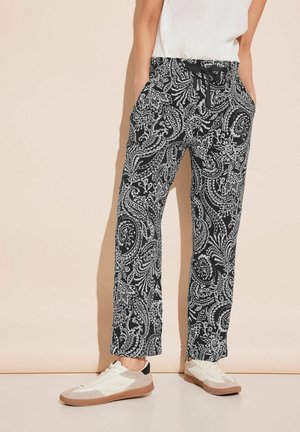 Personne portant un pantalon à motif cachemire noir et blanc avec taille élastique et baskets blanches, debout devant un fond beige.