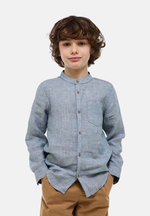 Jeune garçon aux cheveux bouclés portant une chemise bleu clair à boutons et un pantalon marron, debout les mains dans les poches devant un fond uni.