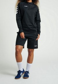 Svart hoodie och shorts av mjukt tyg. Hoodien har vit logotyp och randiga axlar. Blå sportskor och vita strumpor kompletterar outfiten.