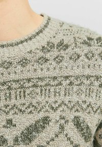 Strickpullover mit runder Ausschnitt, hellbeige Grundfarbe und grünen geometrischen Mustern sowie feiner Textur im gesamten Stoff.