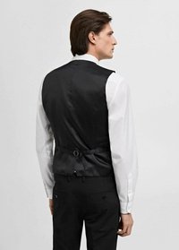Schwarze Weste mit satiniertem Finish, mit einer Schnalle auf der Rückseite, getragen über einem weißen Langarmhemd. Maßgeschneiderter Schnitt.