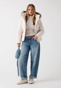 Donzige, lichtroze jas met een capuchon gevoerd met bont, gecombineerd met blauwe wijde jeans. Witte sneakers en een kleine blauwe handtas maken de outfit compleet.