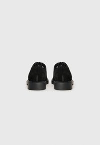 Schwarze Wildlederschuhe mit runder Zehenform und einer flachen Gummisohle, die ein einzigartiges Ausschnittdesign an der Rückseite zur Belüftung aufweisen.