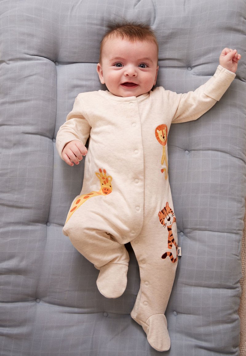 Bébé souriant en grenouillère beige avec des imprimés de lion, girafe et tigre, allongé sur une surface grise rembourrée avec un bras levé.
