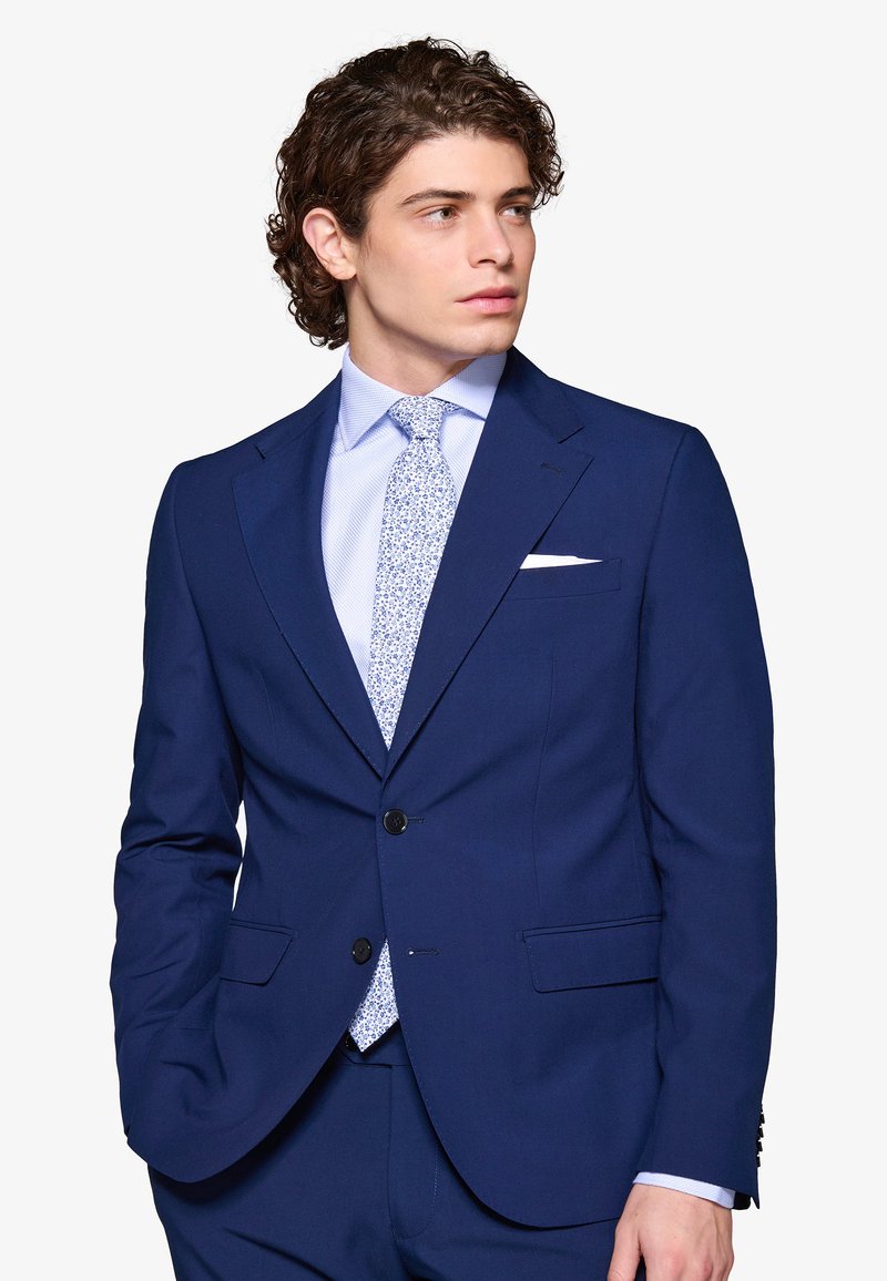 Joven con cabello rizado, vestido con un traje azul oscuro, camisa azul clara y corbata con estampado floral, mirando hacia la derecha.