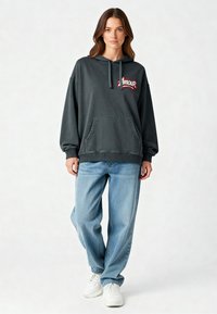 Felpa oversize grigio scuro con tasca frontale e logo "Amour" ricamato in rosso, abbinata a jeans denim azzurri e sneakers bianche.